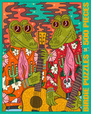 A colorful jigsaw puzzle package front featuring two cartoon alligator-like characters playing guitars, surrounded by flowers, cacti, and psychedelic swirls. The package is labeled 'BIRDIE PUZZLES 500 PIECES' on a light gray background.